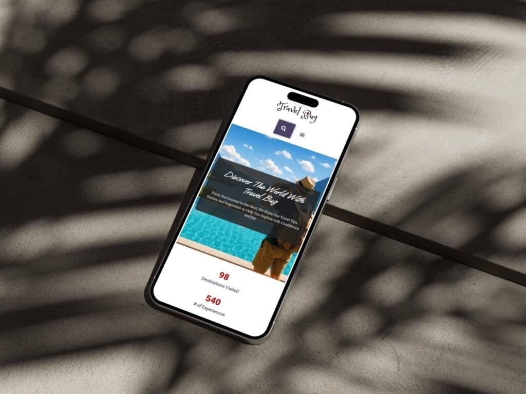 A smartphone displays the TravelBug website, featuring a traveler in a hat overlooking a turquoise pool with the headline “Discover The World With TravelBug.” The phone rests on a textured surface with tropical leaf shadows cast across it, creating a warm, adventurous atmosphere.