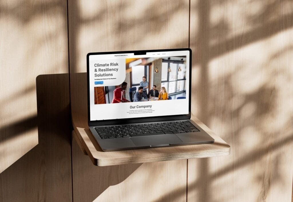 A laptop mounted on a small wooden wall shelf displays the Phil Michele Consulting website, featuring the headline “Climate Risk & Resiliency Solutions” with a team meeting image. Sunlight filters through, casting soft shadows across the light wood wall in the background.