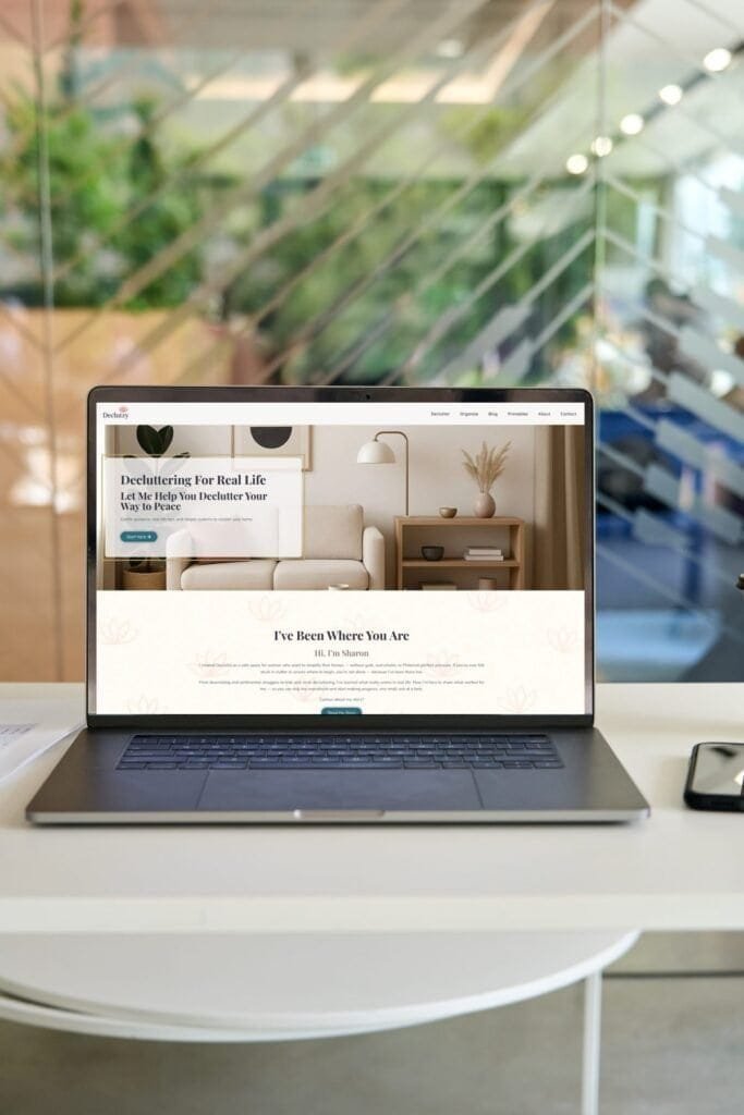 Laptop on an office desk displaying the redesigned Declutzy homepage with a minimalist beige living room scene, promoting decluttering services, with blurred office background.